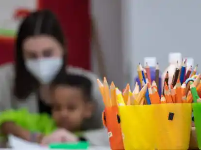 Eine Erzieherin, die eine medizinische Maske tr&auml;gt, sitzt mit einem Kind in einer Kita hinter Bechern mit Buntstiften. Der Landkreis Aurich will die Tr&auml;gerschaft f&uuml;r Kitas und Krippen &uuml;bernehmen.
