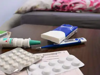 Ein Fieberthermometer, Medikamente und eine &auml;rztliche Krankmeldung liegen auf einem Nachttisch. Viele Menschen sorgen sich um ihre Gesundheit nach &uuml;berstandenen Corona-Infektionen.