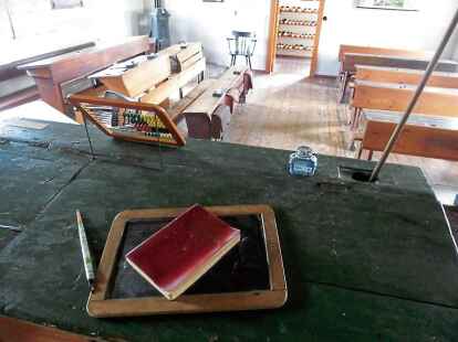 Blick in eine alte Schulstube, in der die Sch&uuml;lerinnen und Sch&uuml;ler auf Holzb&auml;nken eng beieinander sa&szlig;en und in der mit einfachen Rechenmaschinen gerechnet wurde.