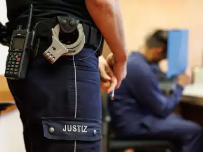 Ein Justizbeamter bewacht den Angeklagten, der sein Gesicht hinter einem Aktendeckel verbirgt.