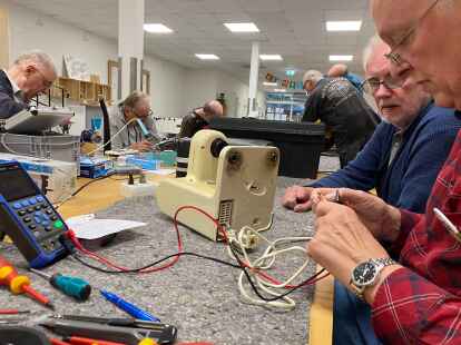 Claus F&ouml;ste (rechts) beim Versuch, eine alte Bosch-K&uuml;chenmaschine wieder zum Laufen zu bringen. Blick auf die lange Werkbank: Werkzeug, Messger&auml;te und Ersatzmaterial so weit das Auge reicht.