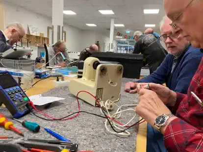 Claus F&ouml;ste (rechts) beim Versuch, eine alte Bosch-K&uuml;chenmaschine wieder zum Laufen zu bringen. Blick auf die lange Werkbank: Werkzeug, Messger&auml;te und Ersatzmaterial so weit das Auge reicht.