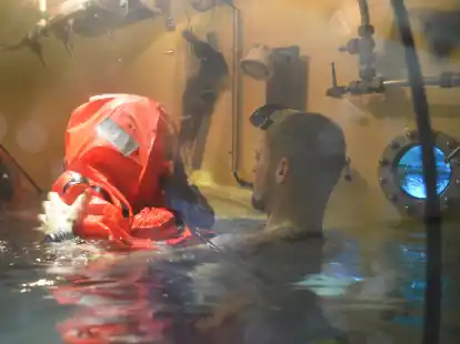 Ein U-Boot-Fahrer im Rettungsanzug mit Ausbilder Konrad in der Schleuse. Das Schott, durch das er in den Tieftauchtopf gelangt, liegt jetzt unter Wasser. Durch das kleine Fenster rechts sieht man in den Tieftauchtopf, in dem der Mann aufsteigen wird.