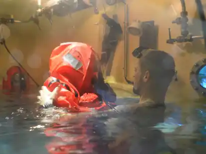Ein U-Boot-Fahrer im Rettungsanzug mit Ausbilder Konrad in der Schleuse.  Das Schott, durch das er in den Tieftauchtopf gelangt, liegt jetzt unter Wasser. Durch das  kleine Fenster rechts sieht man in den Tieftauchtopf, in dem der Mann aufsteigen wird.