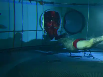 Ein U-Boot-Fahrer im Rettungsanzug sitzt in dem Schott, das die Schleuse mit  dem Tieftauchtopf verbindet. Ein Ausbilder in Badehose taucht zu ihm. Er hilft dem U-Boot-Fahrer zusammen mit weiteren Kameraden bei der Vorbereitung auf den Aufstieg zur  Oberfläche.