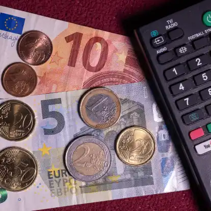 Die Summe von 18,94 Euro liegt auf einem Tisch neben der Fernbedienung f&uuml;r einen Fernseher. Der Rundfunkbeitrag sollte urspr&uuml;nglich ab 2025 auf 18,94 Euro steigen.
